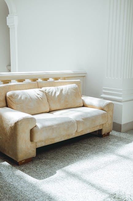 A beige fabric upholstered sofa with plush cushions, positioned against a white wall with decorative molding in a well-lit living room. The light grey carpeted floor appears clean and free of stains, with natural light casting soft shadows across the surface. The setting suggests a neat and maintained space, highlighting the importance of surface cleaning and furniture sanitisation. This image exemplifies professional deep cleaning practices, such as upholstery cleaning, which [COMPANY_NAME] offers to ensure hygiene and freshness in domestic environments, as seen in the context of South Kensington's exhibition road upholstery cleaning and stain removal SW7 services.