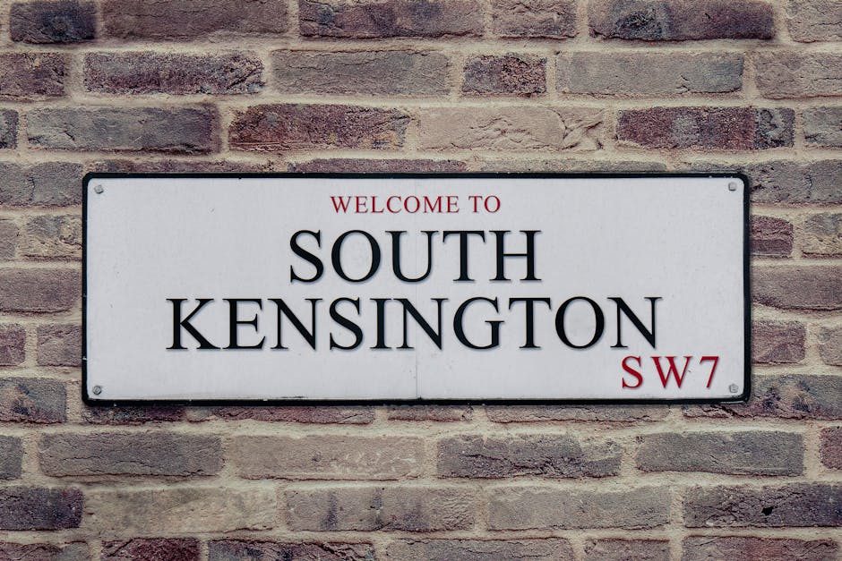 A rectangular metal street sign mounted on a red brick wall, displaying the text 'WELCOME TO SOUTH KENSINGTON SW7' in black and red lettering. The sign is positioned at eye level, with the brick surface showing a mix of light and dark shades and a rough texture. The wall consists of uniformly laid bricks with mortar lines, and the lighting appears natural, illuminating the sign and brickwork clearly. This setting suggests a typical urban environment in South Kensington, emphasizing the importance of surface cleaning and maintenance for safety and aesthetic appeal, as handled by southkensingtoncarpetcleaners.com through comprehensive domestic and commercial cleaning services.