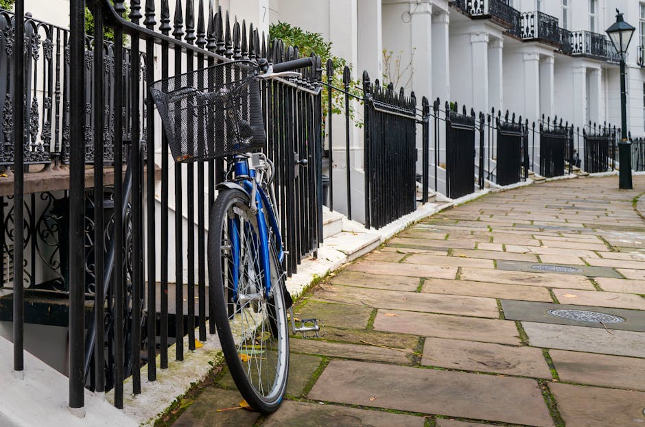 A black wrought iron fence runs alongside a stone-paved sidewalk in South Kensington, with a blue bicycle parked and secured to the fence using a black lock. The sidewalk consists of rectangular stone slabs with some moss or grass growing between them, and the scene is illuminated by natural daylight. In the background, white terraced buildings with black railings on the balconies and black lantern-style wall lights are visible, contributing to a classic London residential aesthetic. The focus on cleanliness aligns with surface and pathway maintenance, an aspect emphasized by South Kensington carpet cleaners for ensuring hygienic and sparkling environments. The overall scene showcases a tidy outdoor area with well-maintained surfaces, suitable for home or commercial property upkeep, reflecting the expertise of Southkensingtoncarpetcleaners.com in deep cleaning and surface sanitation.