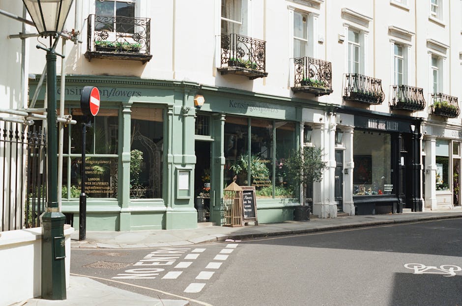 Exterior view of a row of historic storefront buildings in South Kensington, London, with large glass display windows showcasing plants and merchandise. The buildings have ornate iron balconies and white-painted facades. A small chalkboard sign stands outside one shop, and the street is paved with asphalt, featuring a bicycle lane with painted symbols. Natural daylight illuminates the scene, emphasizing the clean and well-maintained appearance of the storefronts. This image highlights the aesthetic appeal of the area, relevant to surface cleaning and maintenance services provided by southkensingtoncarpetcleaners.com for commercial properties in the SW7 area near South Kensington station.
